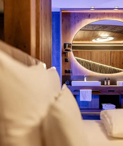 Blick vom Bett auf den offenen Waschtisch mit beleuchtetem Rundspiegel und Doppelwaschbecken in einer Suite des Hotel Kronblick
